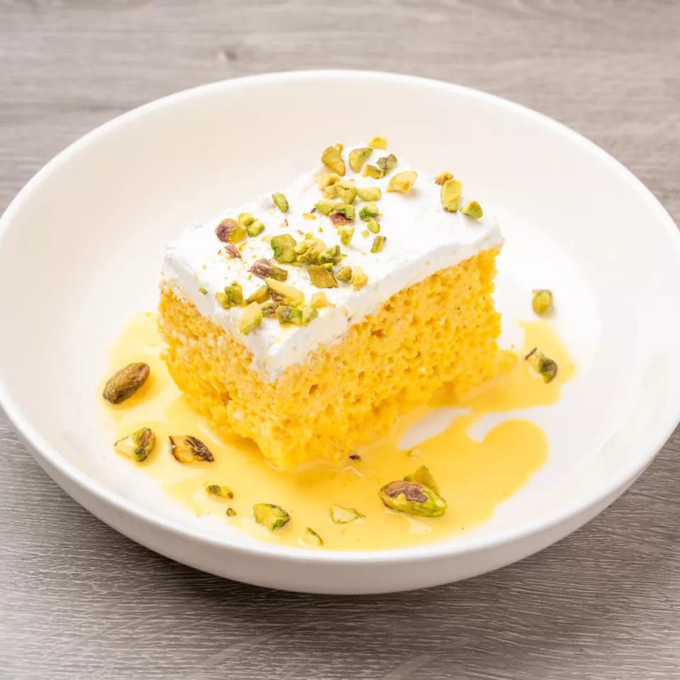 Slice of yellow cake with white frosting, drizzled with yellow sauce and topped with pistachios.