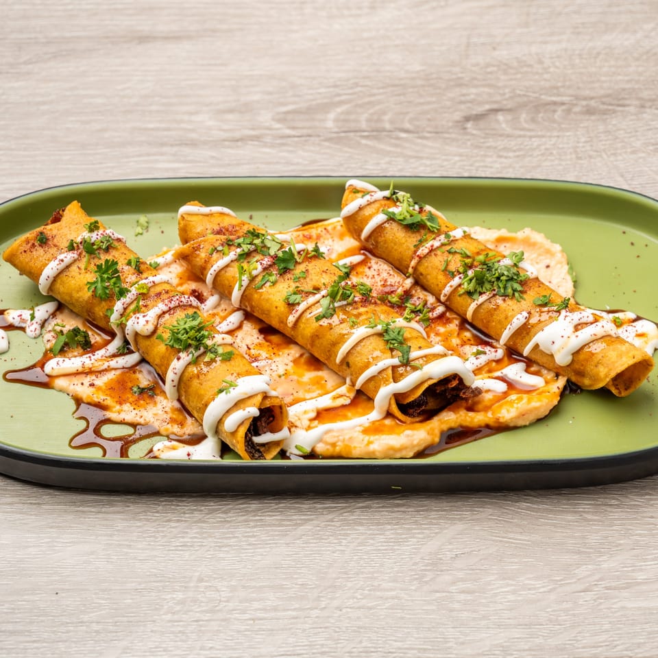 Four golden taquitos drizzled with white sauce, cilantro, and spices on a green plate.