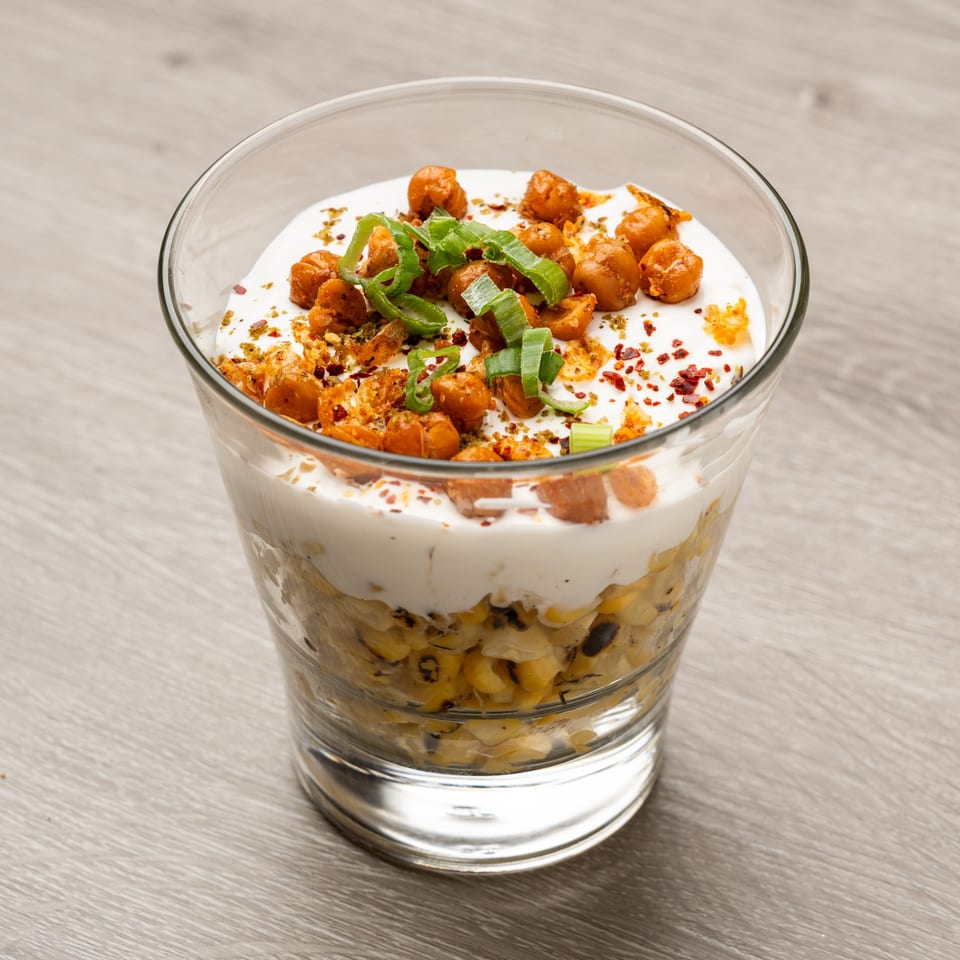 Elote-style corn salad in a glass, topped with white sauce, crispy chickpeas, and green onions.