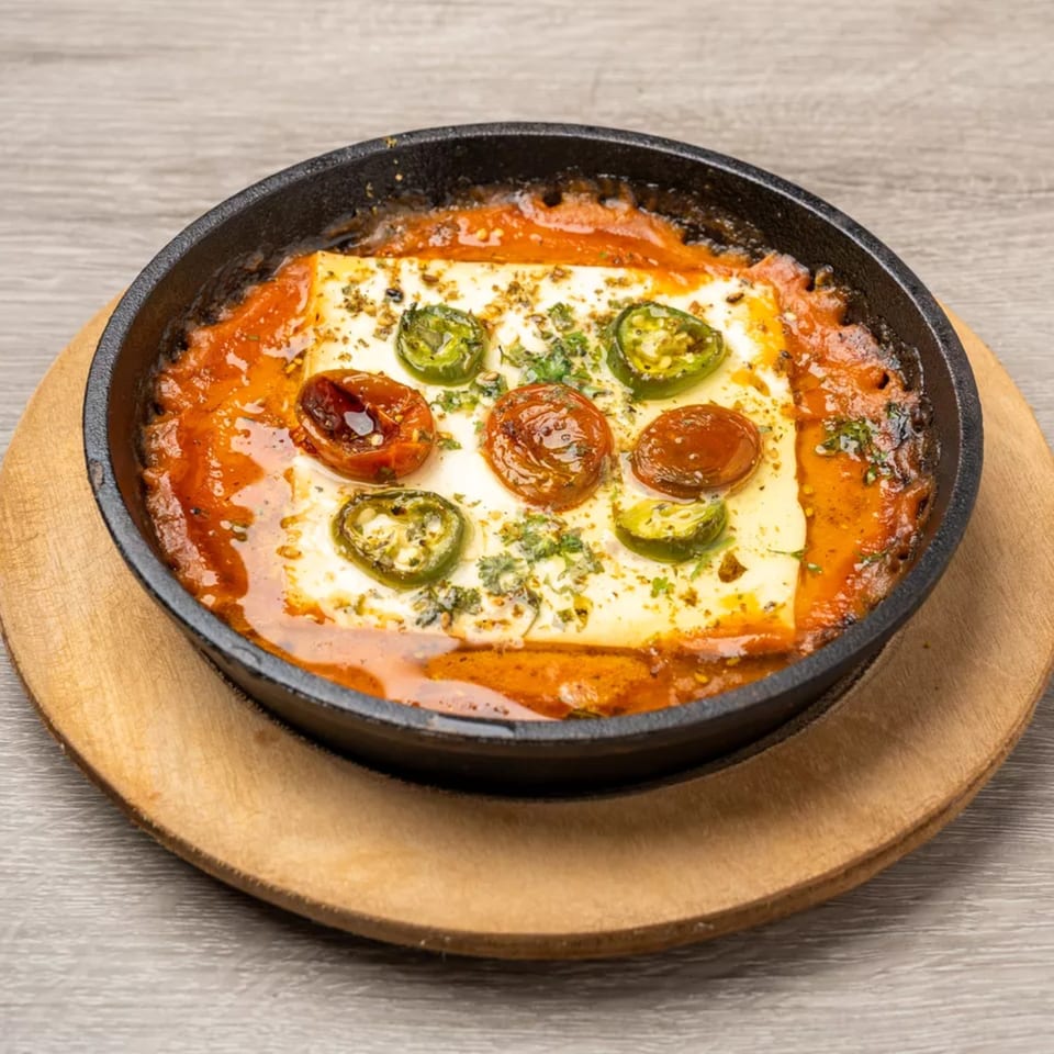 Baked feta cheese in tomato sauce with jalapeños and herbs, served in a black skillet on a wooden board.
