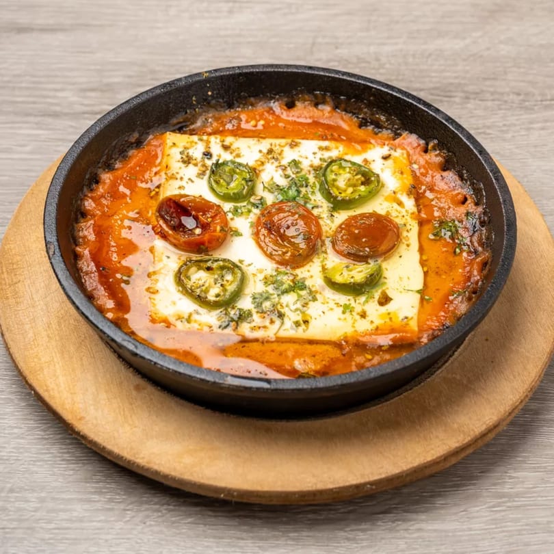 Baked feta cheese in tomato sauce with jalapeños and herbs, served in a black skillet on a wooden board.