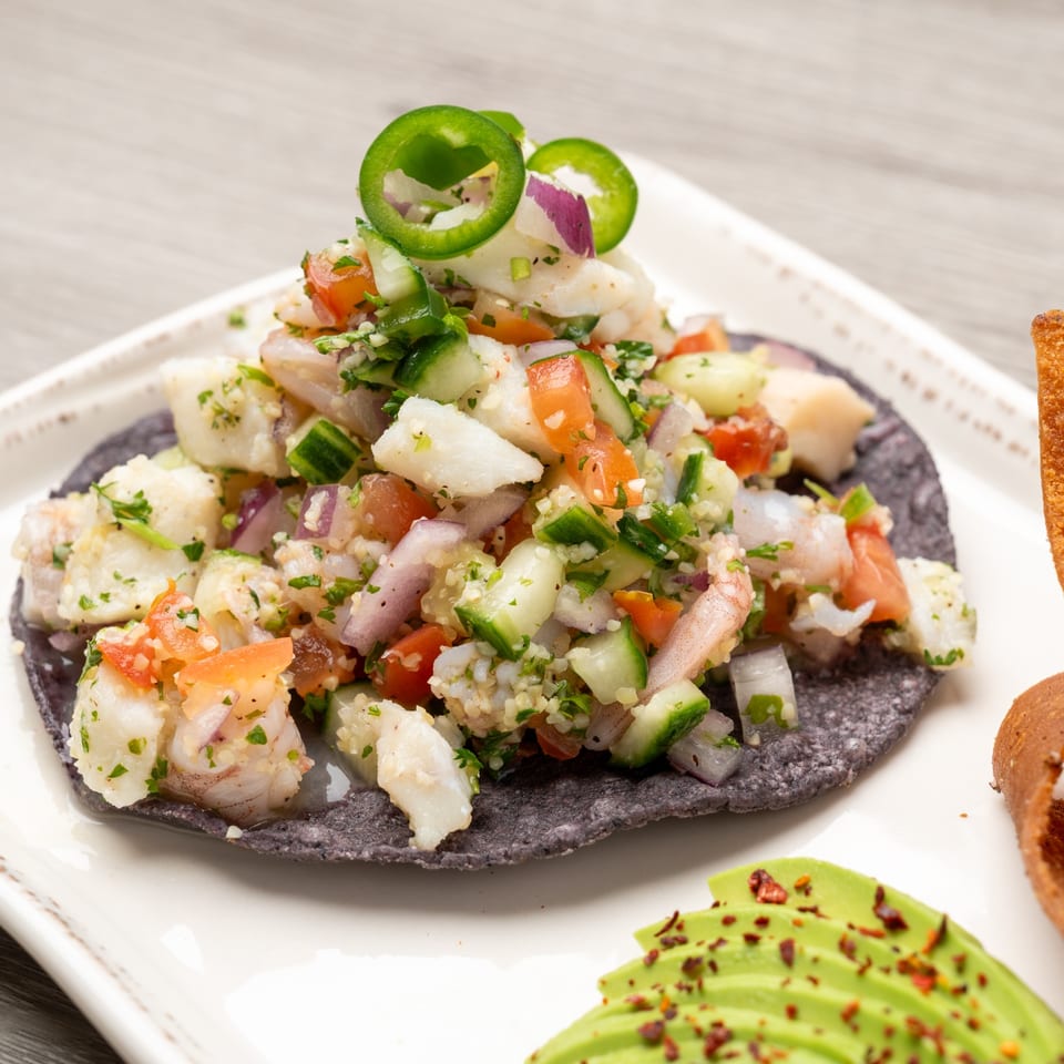 Ceviche on a blue corn tostada, topped with jalapeños, served with avocado slices.