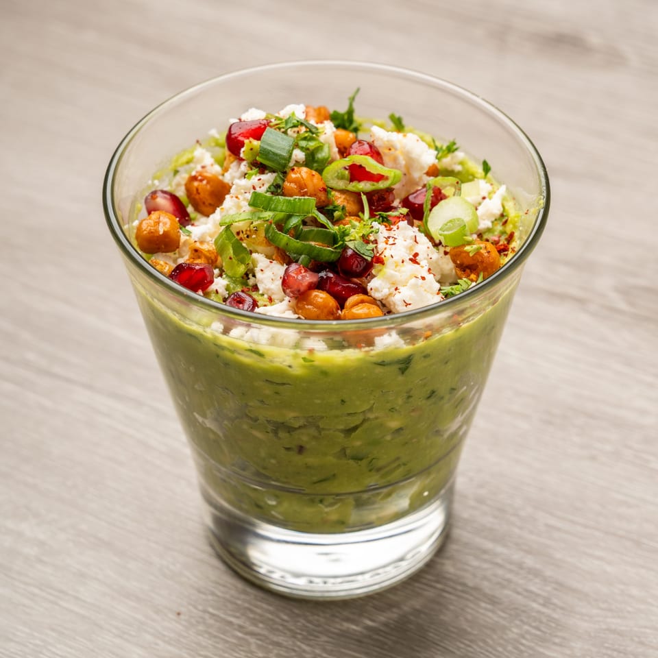 Guacamole in a glass topped with feta, chickpeas, pomegranate seeds, and green onions.